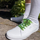 White sneakers with green and yellow laces worn with white socks on a concrete surface.