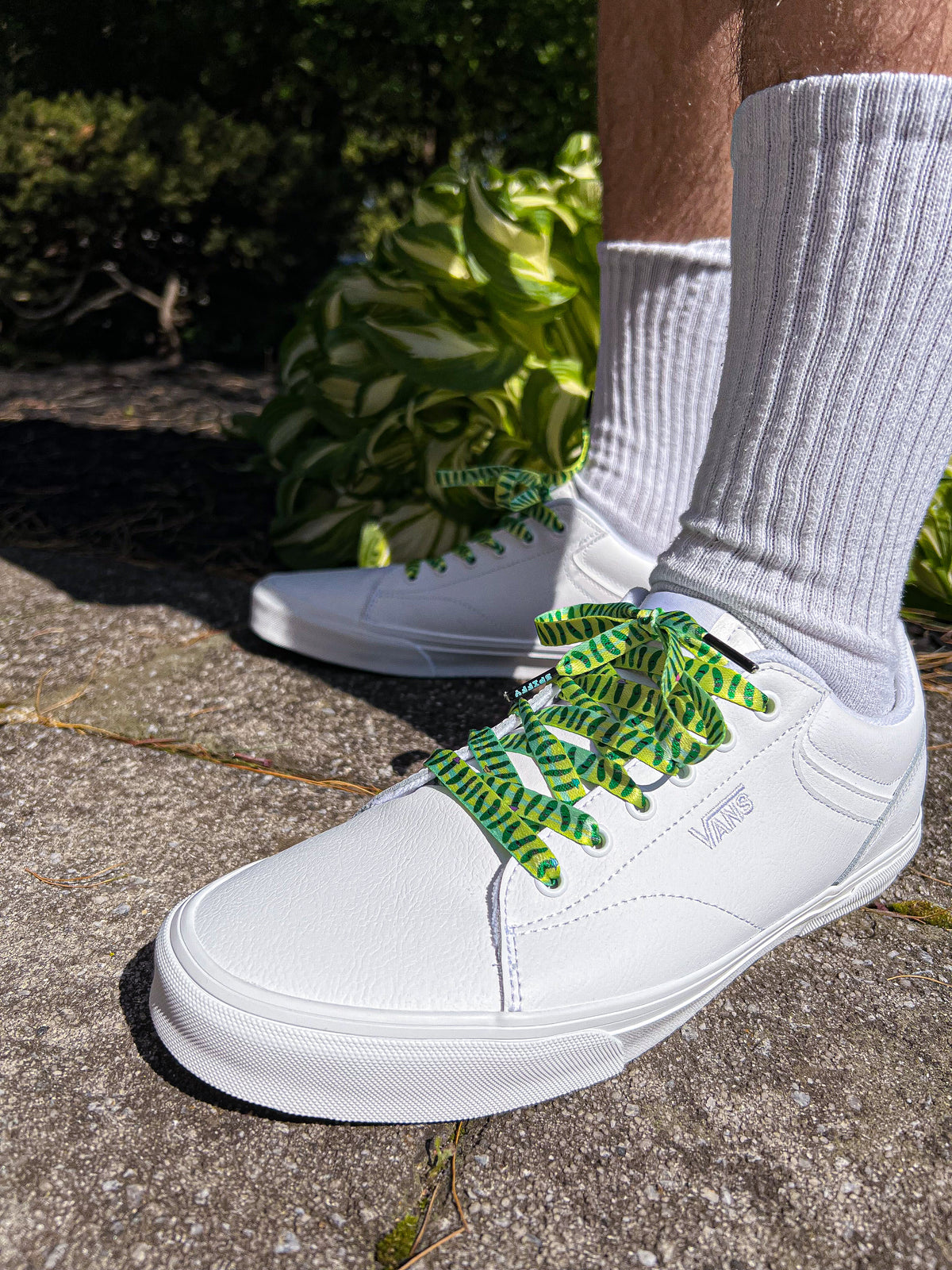 White sneakers with green and yellow laces worn with white socks on a concrete surface.