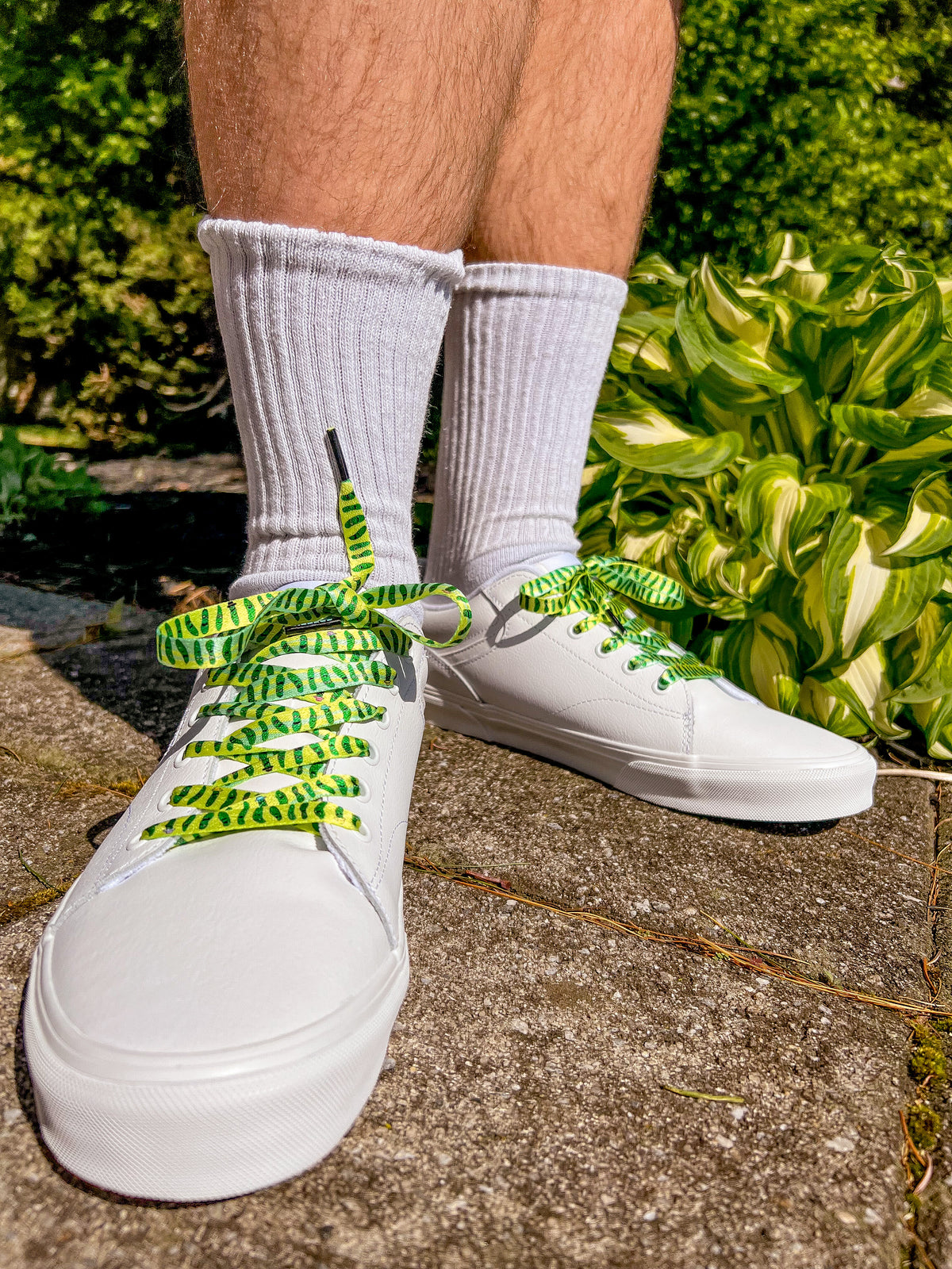 White sneakers with green and yellow pickle laces worn by a person on a concrete surface with greenery in the background.