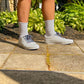 Person wearing light gray sneakers and retro themed zig zag shoelaces on a sidewalk with greenery in the background