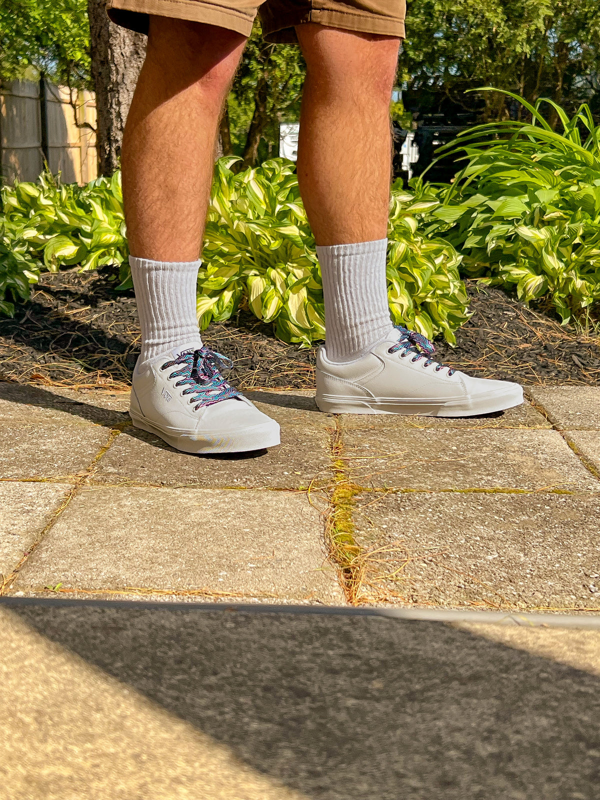 Person wearing light gray sneakers and retro themed zig zag shoelaces on a sidewalk with greenery in the background