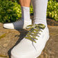 White sneakers with green and yellow psychedelic smiley face patterned laces worn with white socks outdoors.