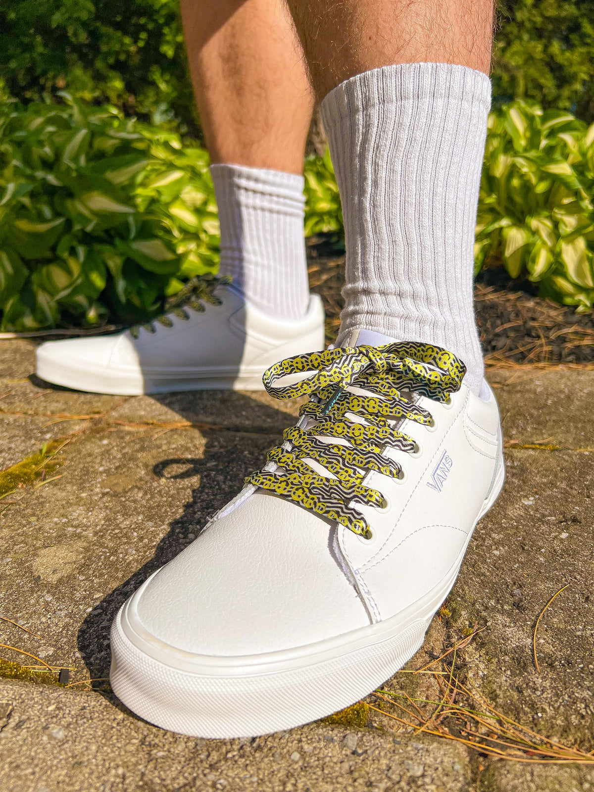 White sneakers with green and yellow psychedelic smiley face patterned laces worn with white socks outdoors.