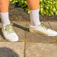 White sneakers with psychedelic smiley face patterned laces worn on a concrete surface with greenery in the background