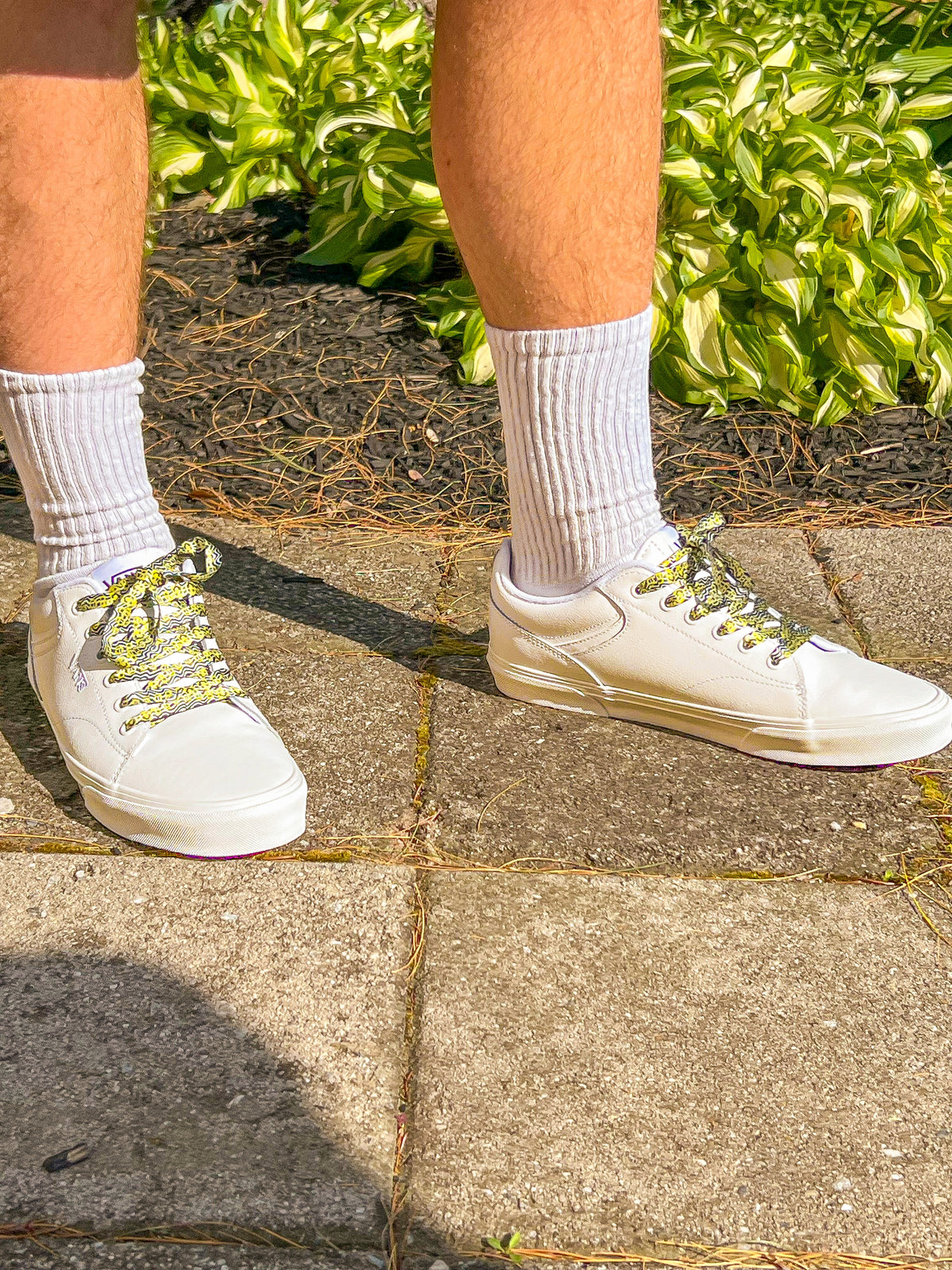 White sneakers with psychedelic smiley face patterned laces worn on a concrete surface with greenery in the background