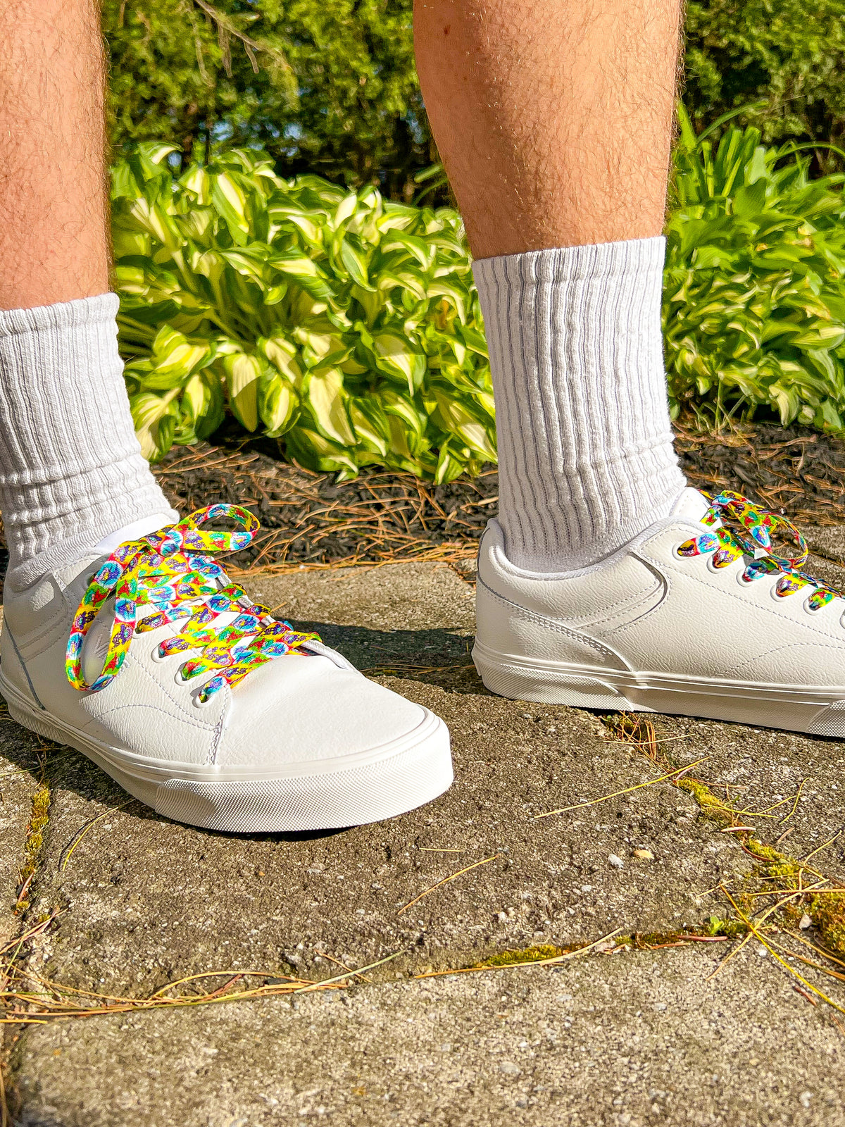 White sneakers with colorful laces worn on a concrete surface with greenery in the background