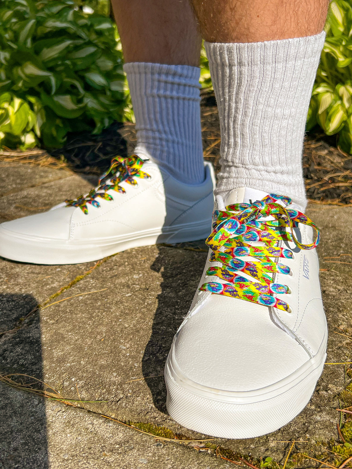 White sneakers with colorful laces worn on a concrete surface with greenery in the background