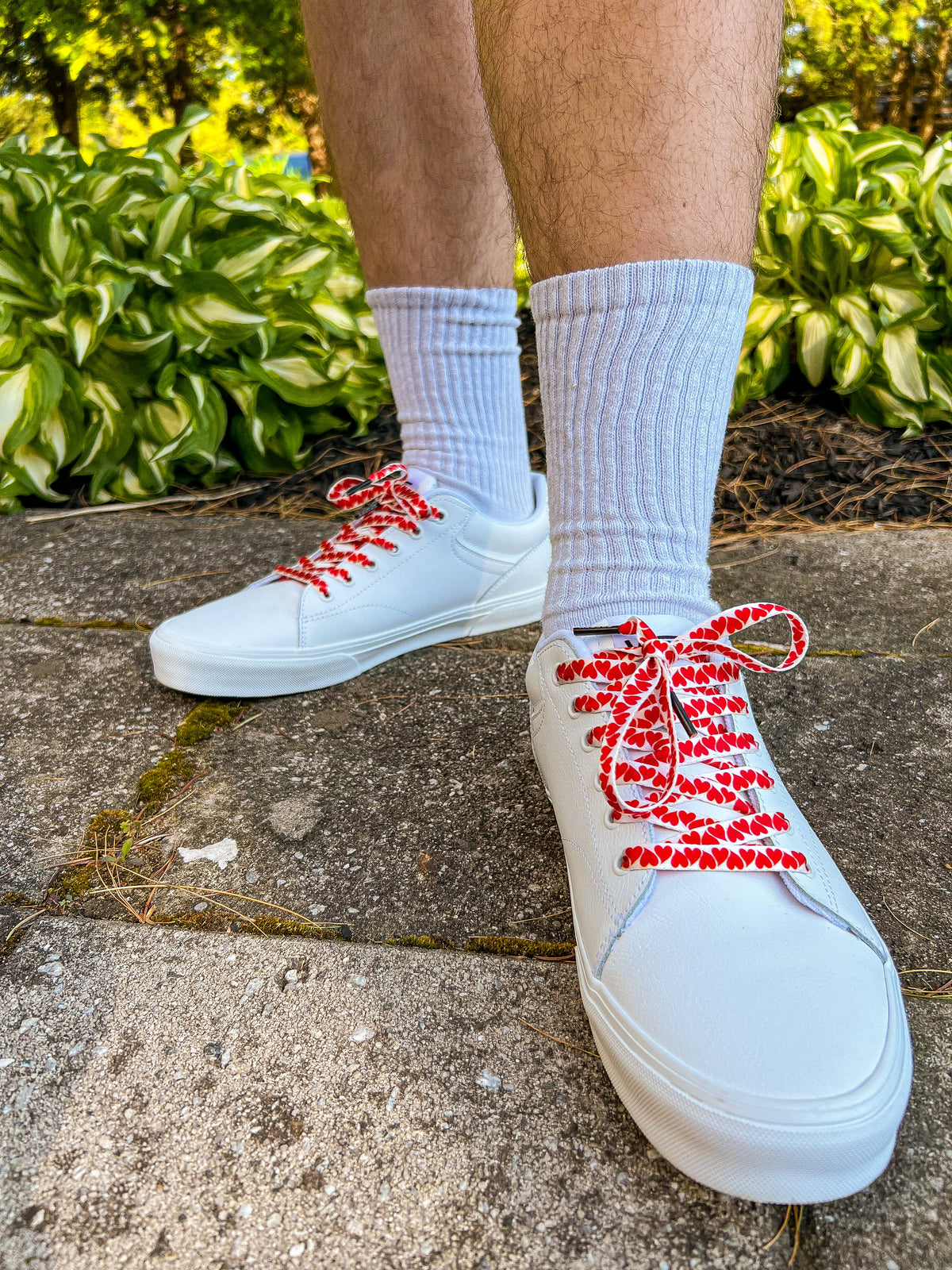White sneakers with colorful heart shoelaces worn on a concrete surface with greenery in the background