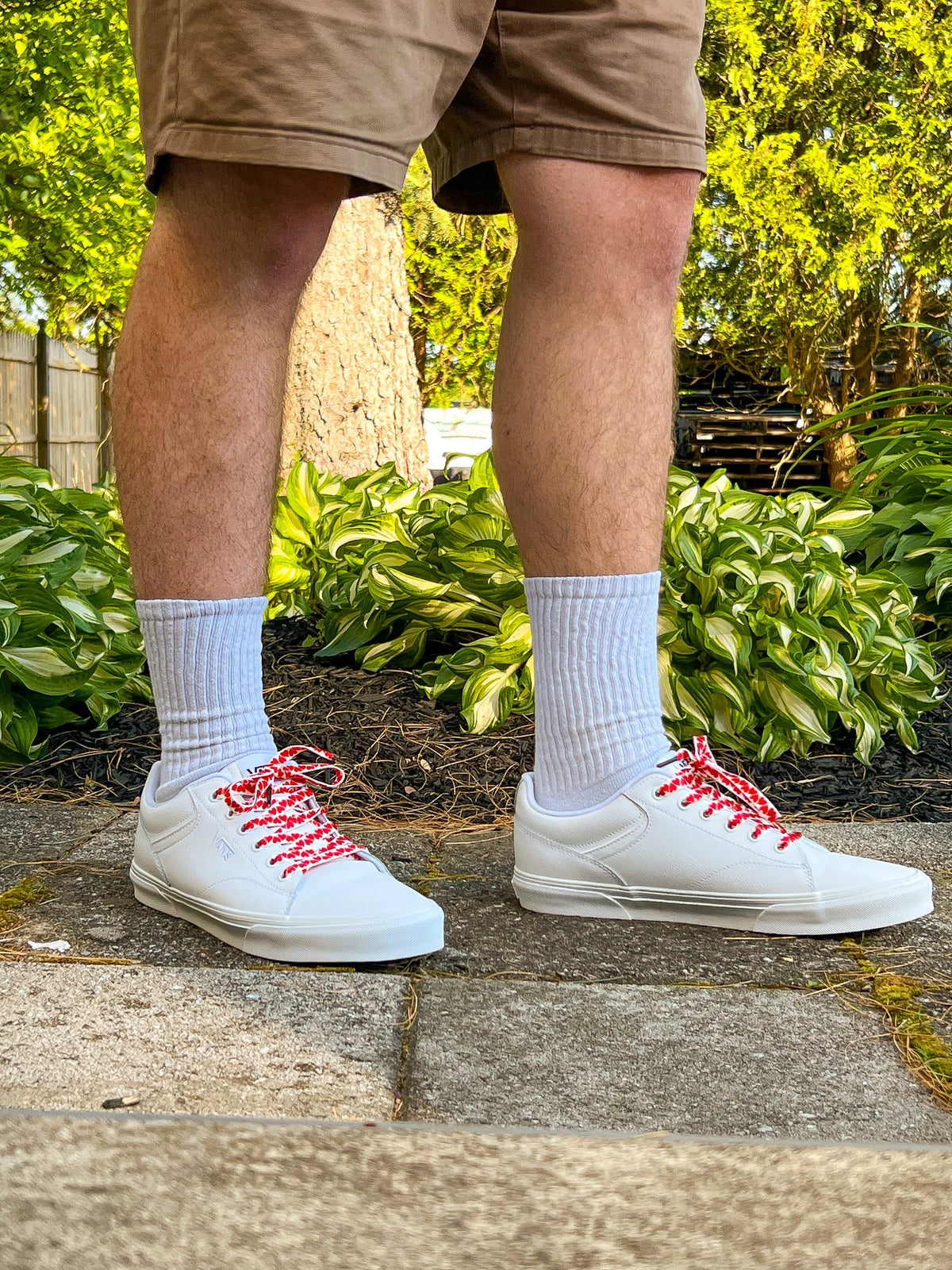 White sneakers with colorful heart shoelaces worn by a person standing on a concrete surface with greenery in the background.