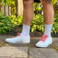 White sneakers with colorful heart shoelaces worn outdoors on a stone path with greenery in the background.
