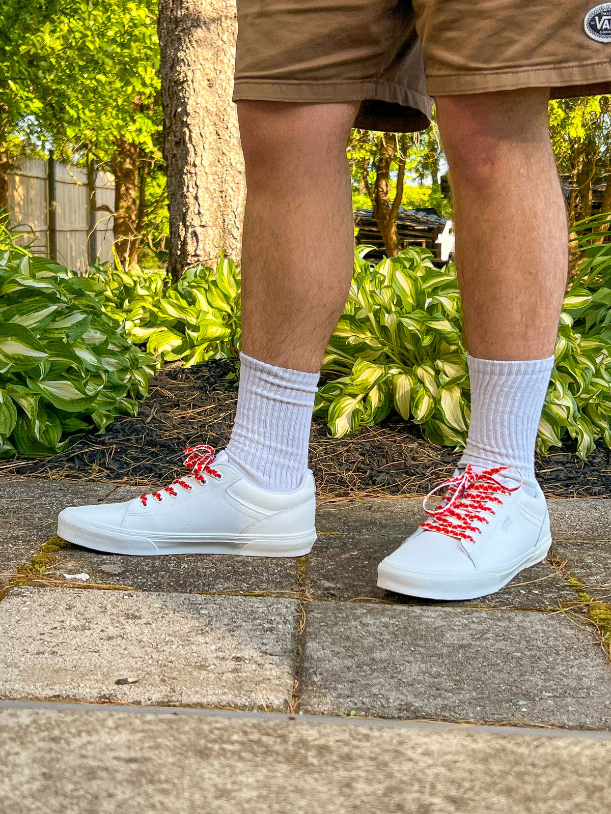 White sneakers with colorful heart shoelaces worn outdoors on a stone path with greenery in the background.