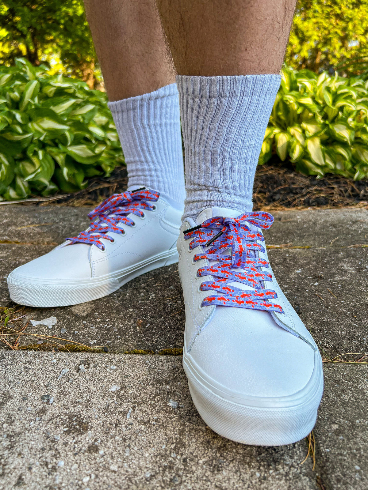 White sneakers with colorful and fun lobster shoelaces worn on a concrete surface with greenery in the background