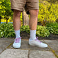 Person wearing white sneakers with colorful and fun lobster shoelaces, beige shorts, and white socks standing on a paved path with greenery in the background.