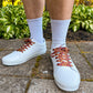 White sneakers with maroon laces worn outdoors on a concrete surface with greenery in the background.