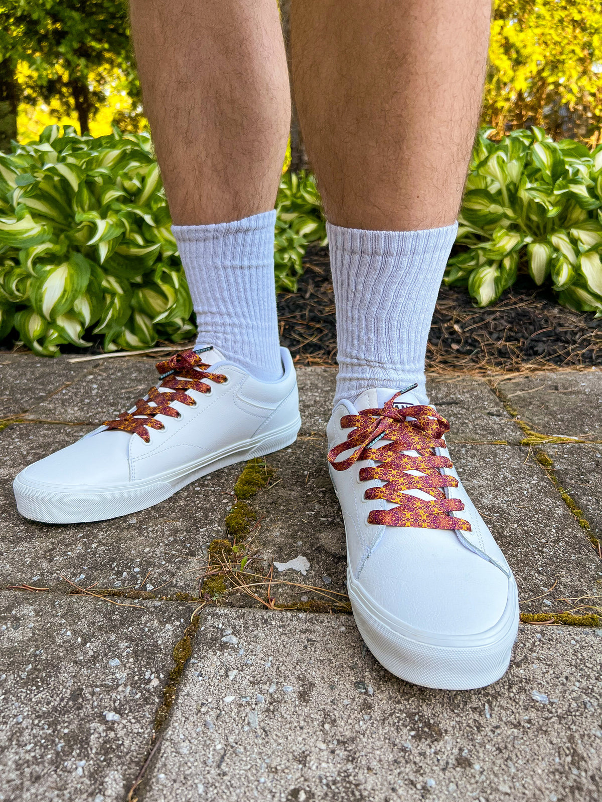White sneakers with maroon laces worn outdoors on a concrete surface with greenery in the background.