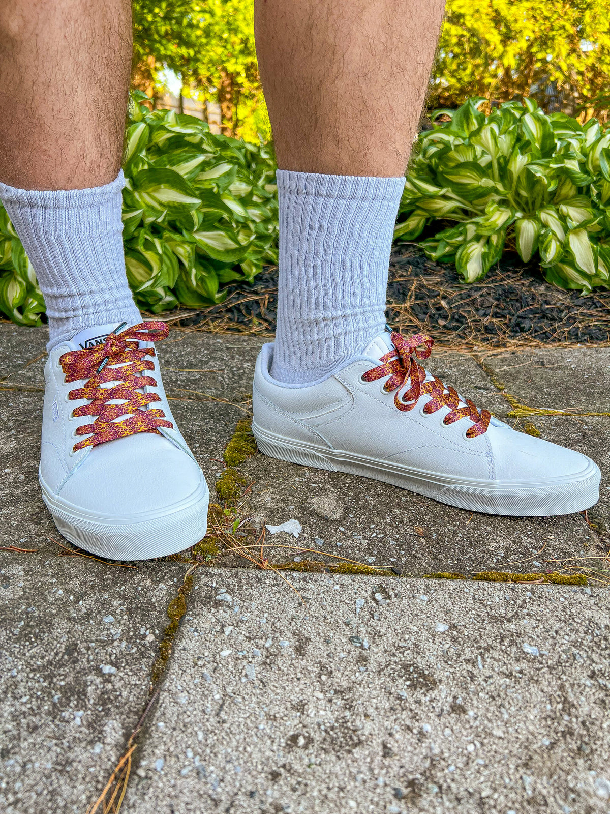 White sneakers with tapestry designed laces worn by a person on a concrete surface with greenery in the background