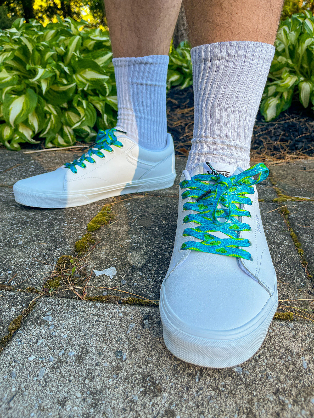 White sneakers with blue laces worn on a concrete surface with greenery in the background