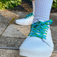 White sneakers with blue laces worn on a concrete surface with greenery in the background
