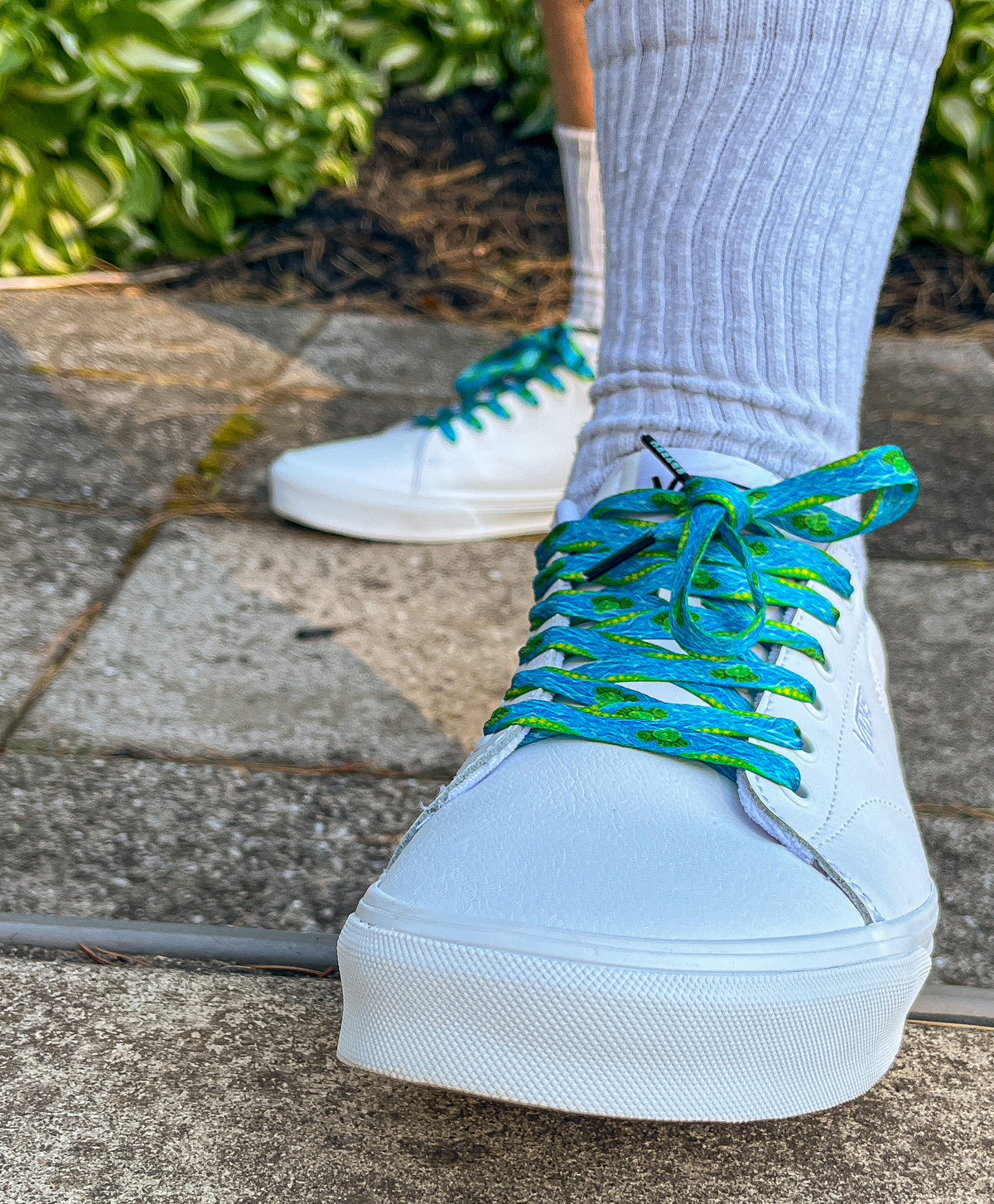 White sneakers with blue laces worn on a concrete surface with greenery in the background
