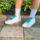 White sneakers with blue laces worn on a stone pavement with green plants in the background