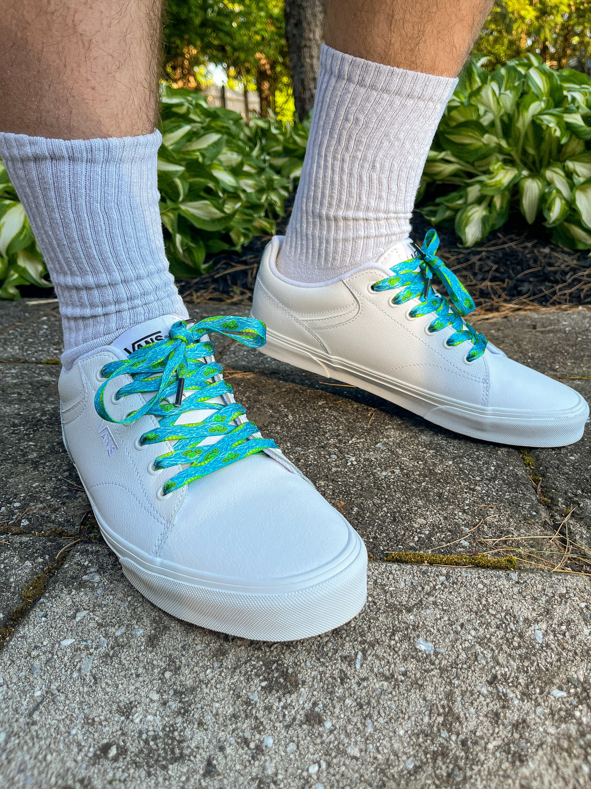 White sneakers with blue laces worn on a concrete surface with greenery in the background