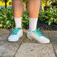 White sneakers with green laces worn on a stone path with greenery in the background