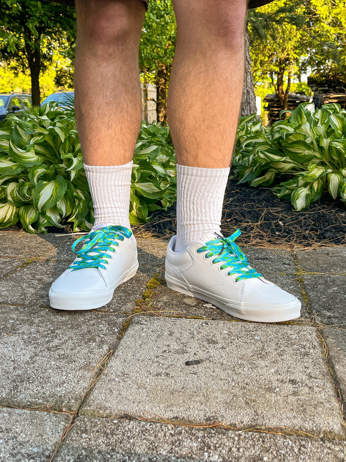 White sneakers with green laces worn on a stone path with greenery in the background