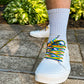 White sneakers with rainbow laces on a stone pavement with green plants in the background
