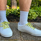 White sneakers with yellow duck themed shoelaces worn on a concrete surface with greenery in the background