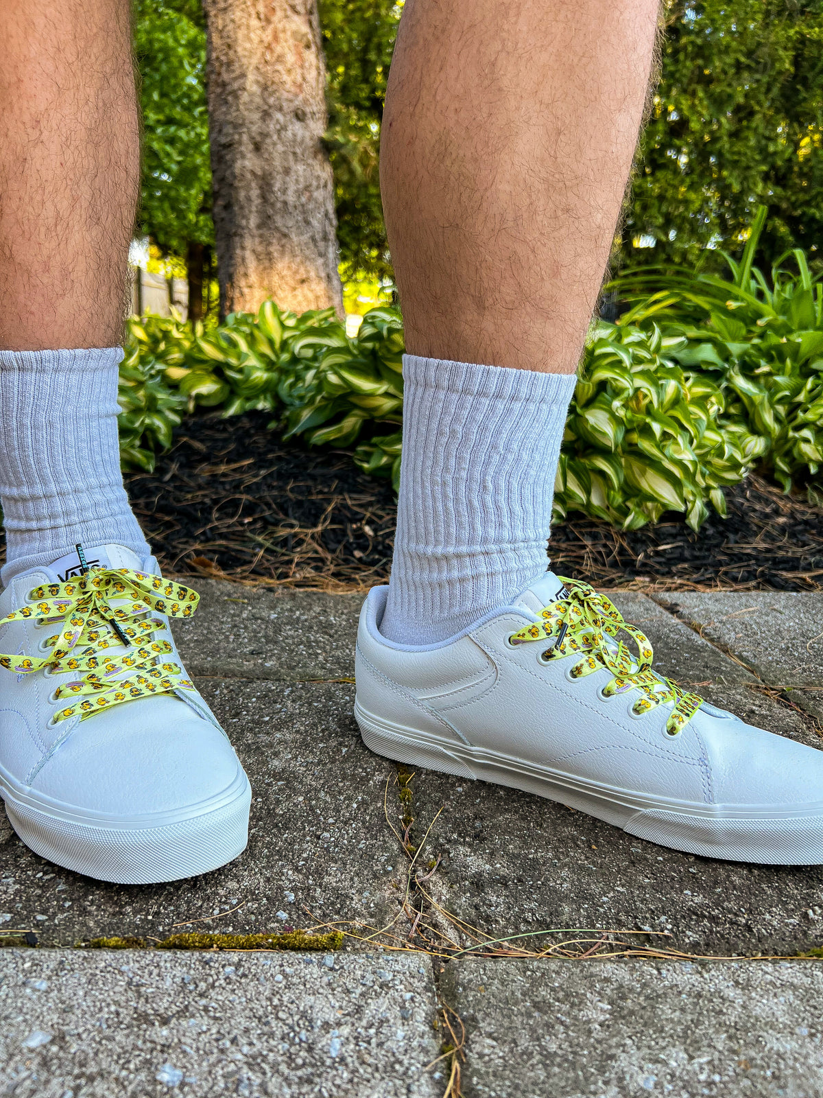 White sneakers with yellow duck themed shoelaces worn on a concrete surface with greenery in the background