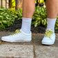 White sneakers with yellow shoelaces with rubber ducks worn on a person's feet, standing on a concrete surface with greenery in the background.