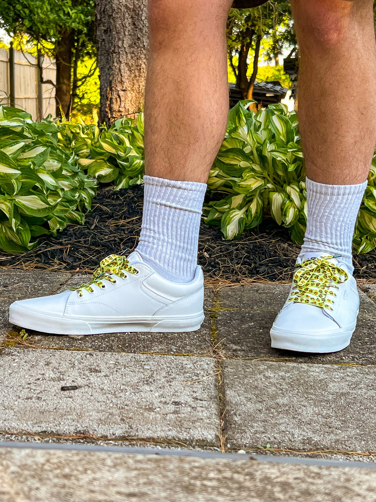 White sneakers with yellow shoelaces with rubber ducks worn on a person's feet, standing on a concrete surface with greenery in the background.