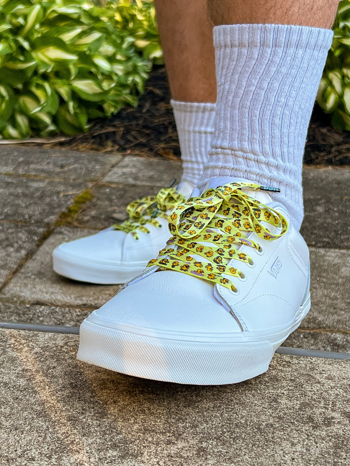 White sneakers with yellow duck themed shoelaces worn with light gray socks on a concrete surface.