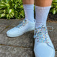 White sneakers with colorful mushroom shoelaces worn on a concrete surface with greenery in the background