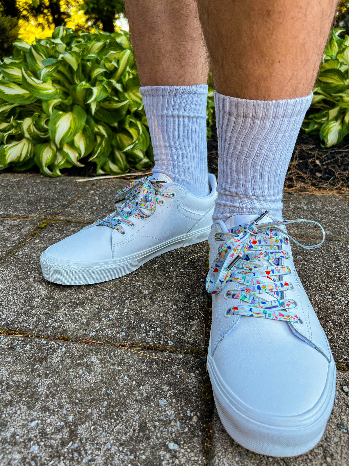 White sneakers with colorful mushroom shoelaces worn on a concrete surface with greenery in the background