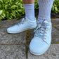 White sneakers with colorful mushroom shoelaces worn on a stone path with greenery in the background