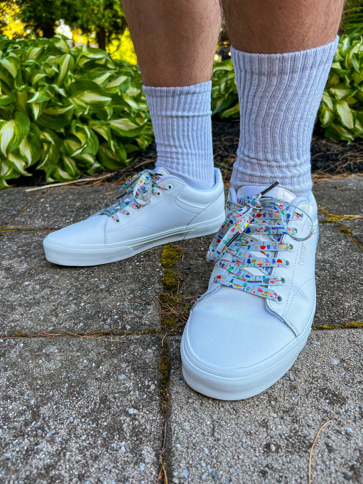 White sneakers with colorful mushroom shoelaces worn on a stone path with greenery in the background