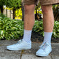 Person wearing white sneakers with colorful mushroom shoelaces, light blue socks, and brown shorts standing on a sidewalk with greenery in the background.