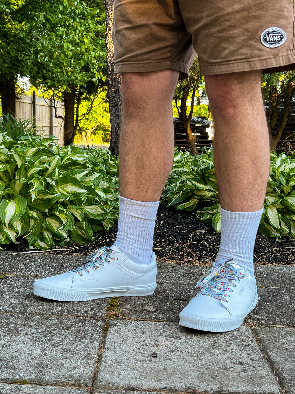Person wearing white sneakers with colorful mushroom shoelaces, light blue socks, and brown shorts standing on a sidewalk with greenery in the background.