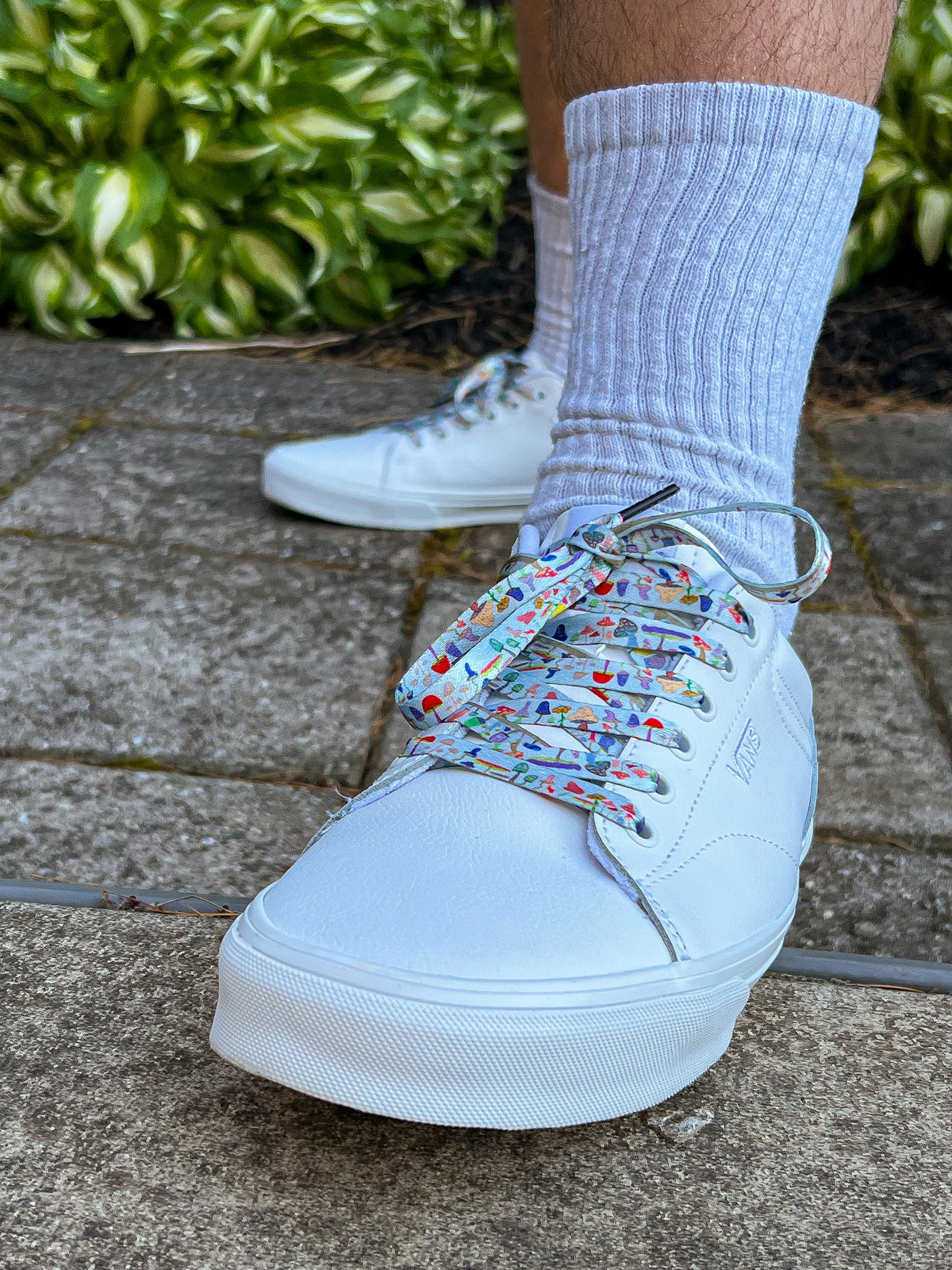 White sneakers with colorful mushroom shoelaces worn on a concrete surface with greenery in the background