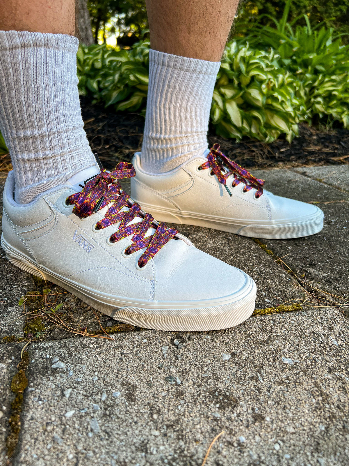 White sneakers with colorful laces worn on a concrete surface with greenery in the background