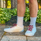 Person wearing white sneakers with maroon laces, light gray socks, and beige shorts on a stone path with greenery in the background.