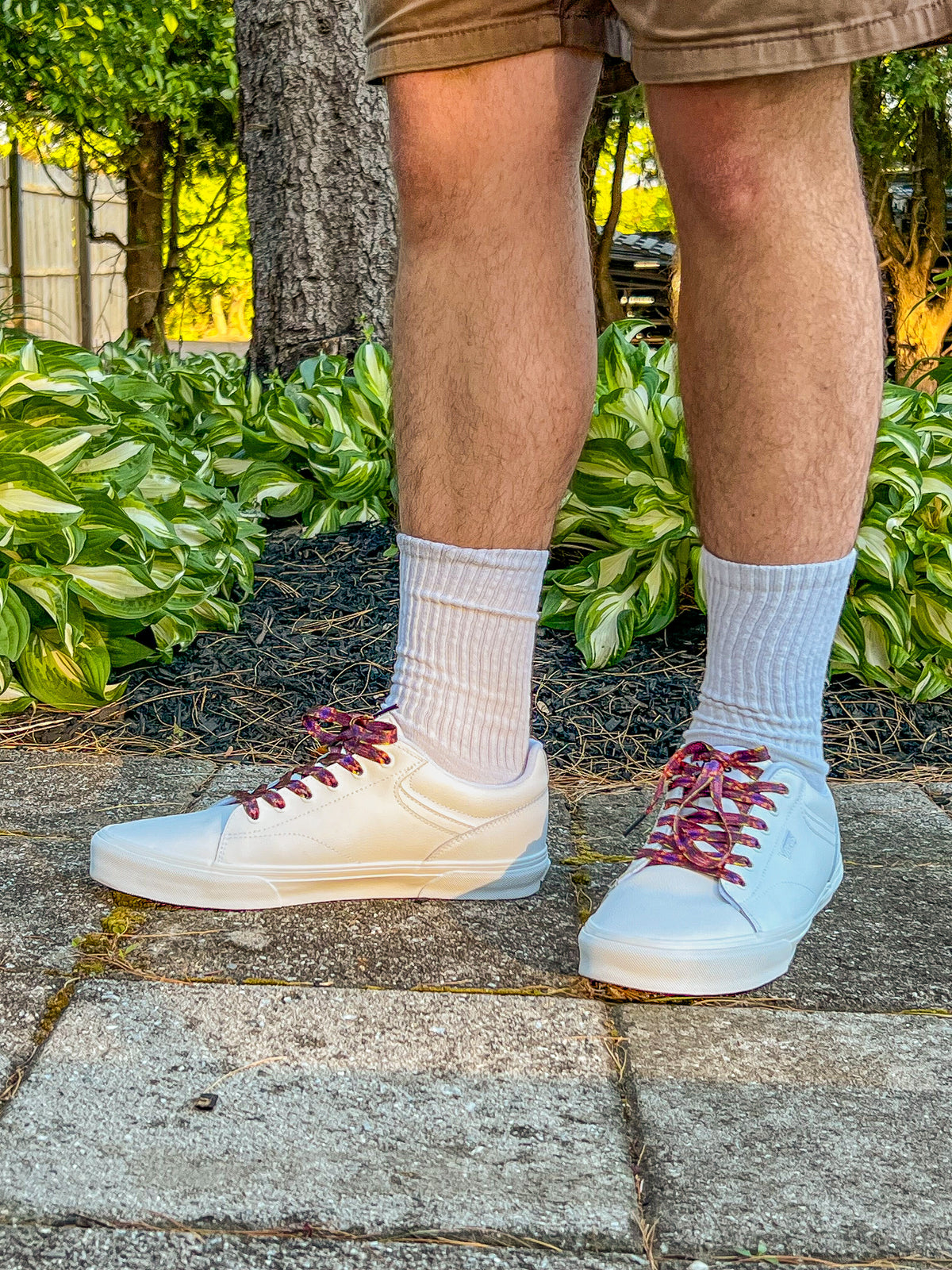 Person wearing white sneakers with maroon laces, light gray socks, and beige shorts on a stone path with greenery in the background.