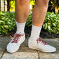 Person wearing white sneakers with Tapestry printed laces on a stone path with greenery in the background