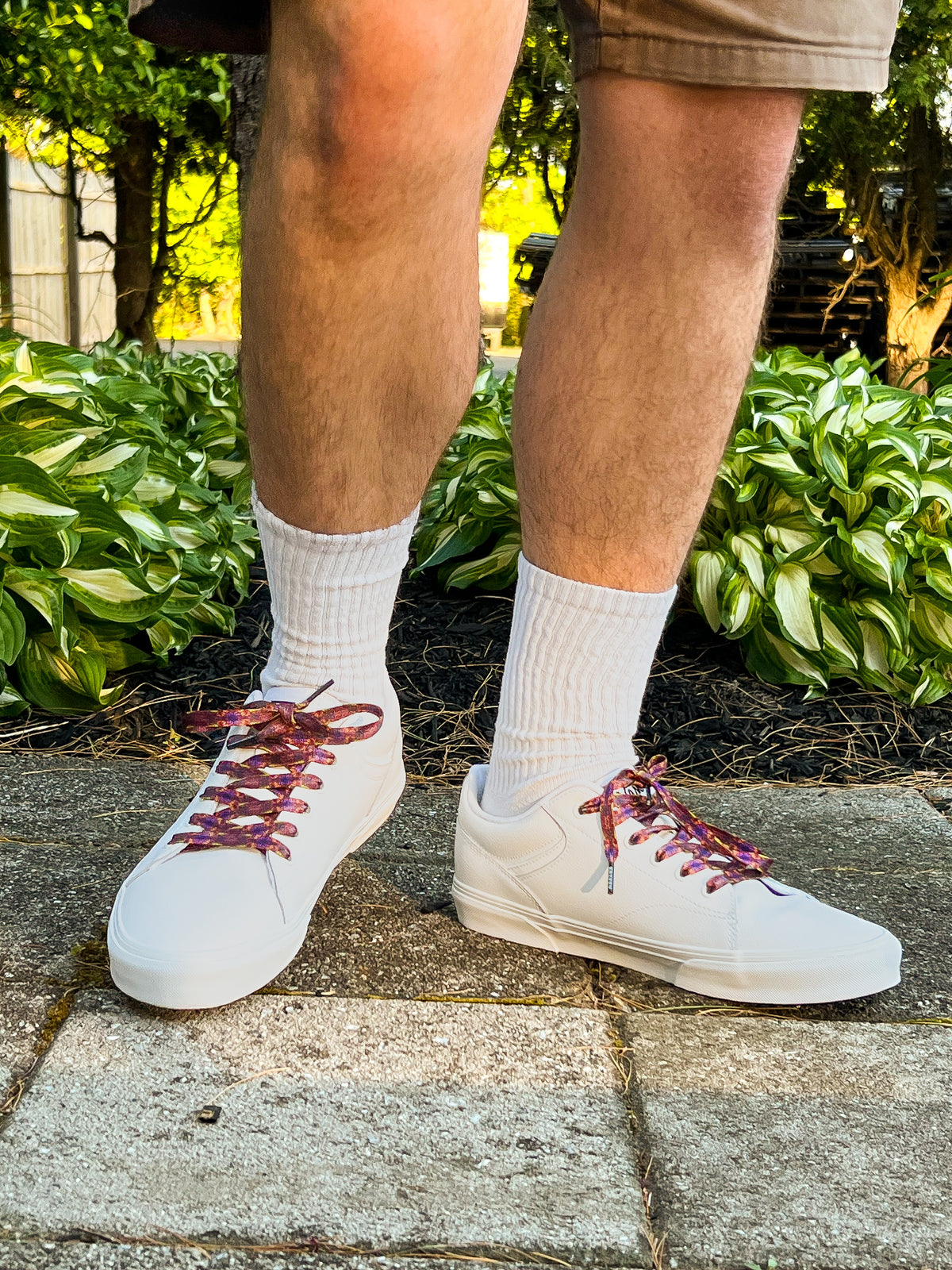 Person wearing white sneakers with Tapestry printed laces on a stone path with greenery in the background