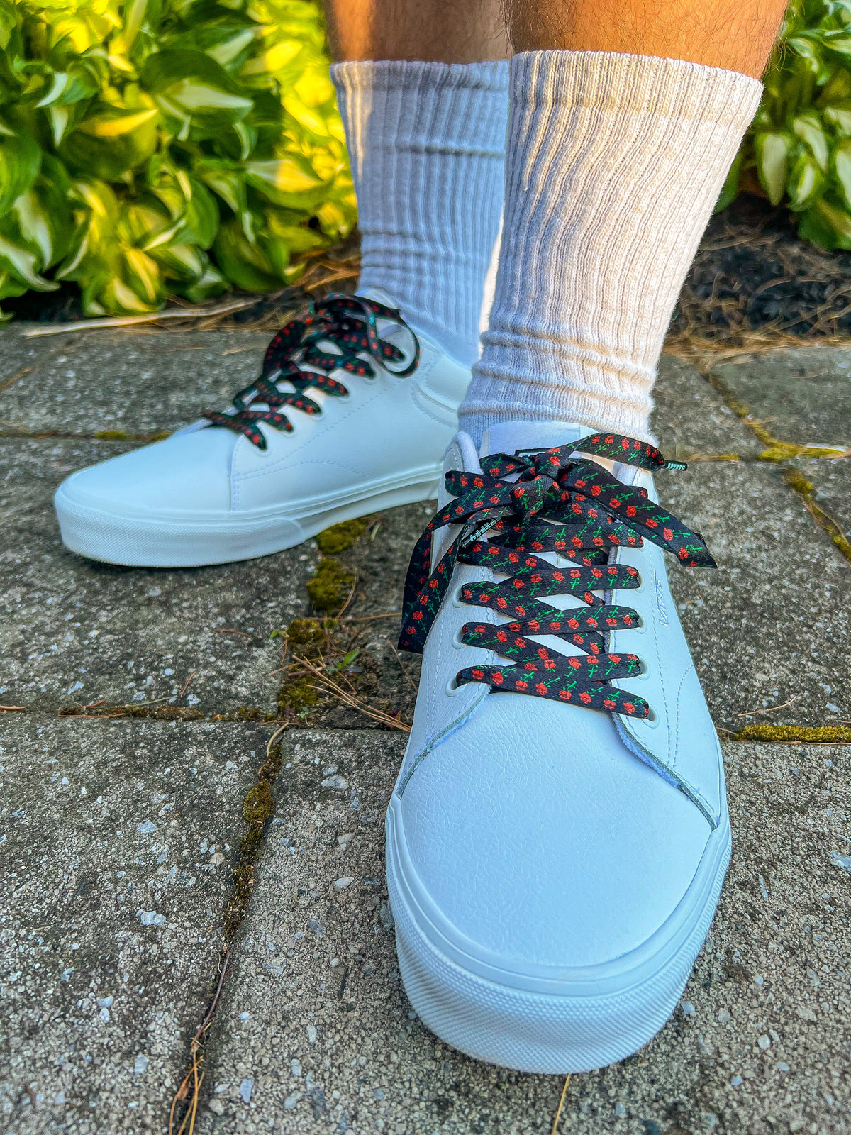 White sneakers with black and red laces worn on a concrete surface with greenery in the background.