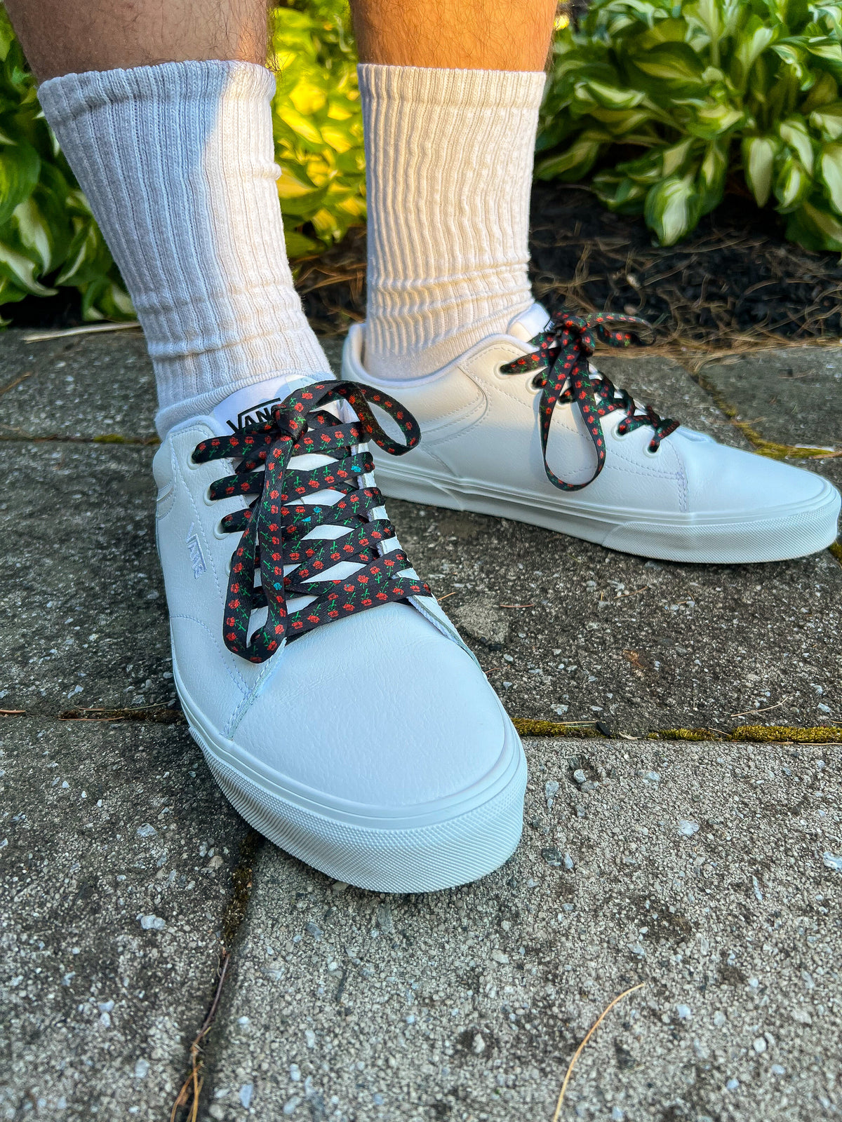 White sneakers with black and red laces worn on a concrete surface with greenery in the background