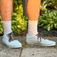 White sneakers with rose shoelaces  worn outdoors on a concrete surface with greenery in the background.