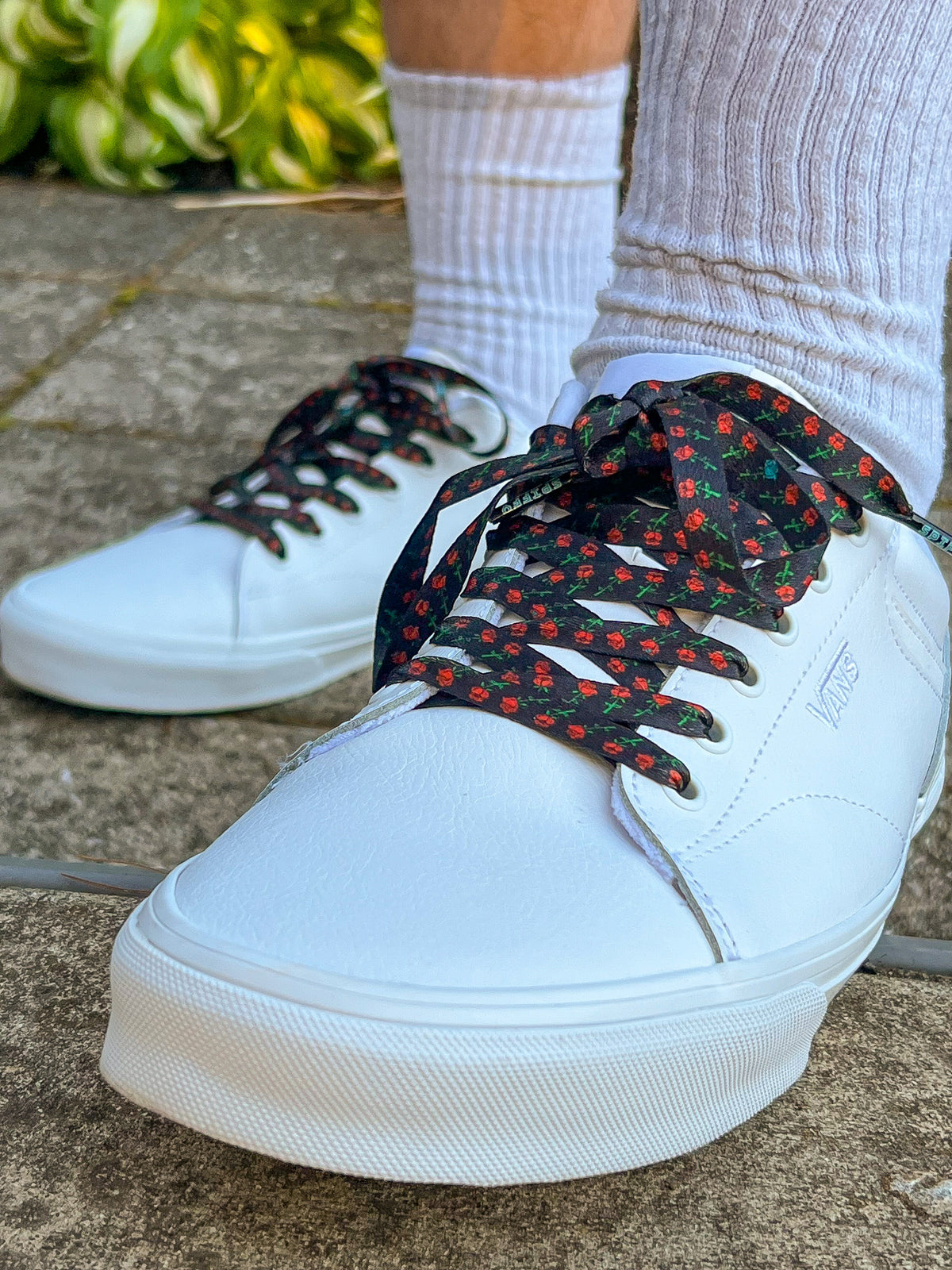White sneakers with black and red rose patterned laces worn on a concrete surface.