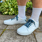 White sneakers with evil eye themed shoelaces worn on a concrete surface with greenery in the background